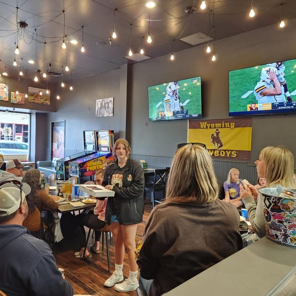A young woman holds a pizza box in a lively restaurant with people, TVs, and pinball machines.