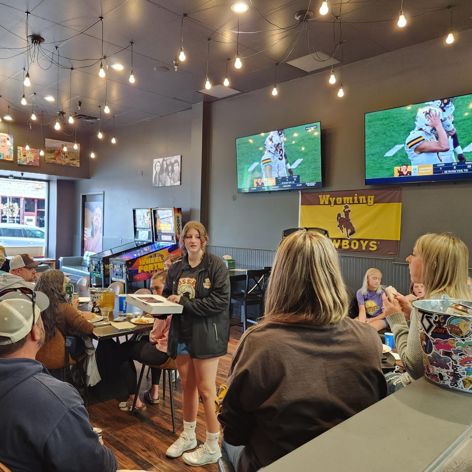A young woman holds a pizza box in a lively restaurant with people, TVs, and pinball machines.